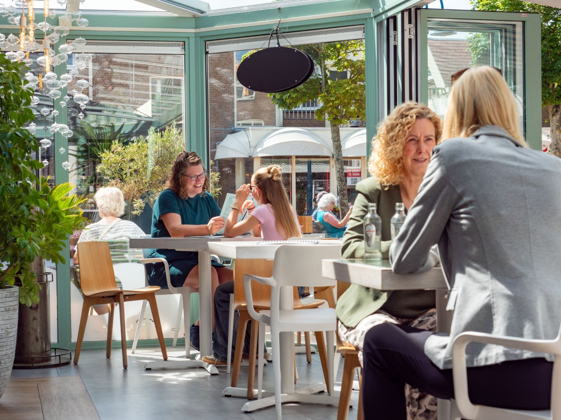 Gasten aan tafel in de lichte serre, met uitzicht door de glazen wanden op de straat