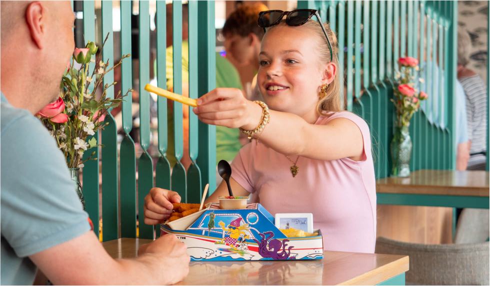 Meisje aan tafel houdt lachend een patatje omhoog, met een kindermenu in een bootje voor zich